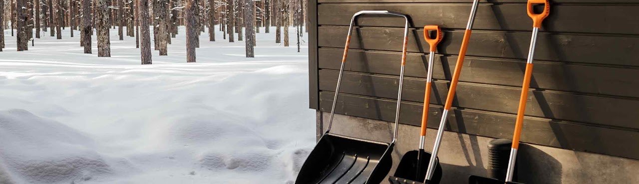 Snöskyfflar och en snöskopa med orange handtag lutade mot en trävägg i snötäckt tallskog
